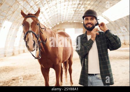 Wearing protective hat and smiling. Young man with a horse is in the hangar. Stock Photo