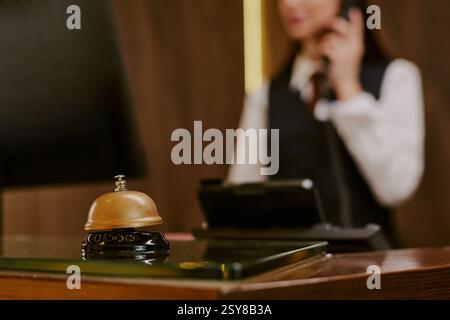 Medium close up of golden bell on polished table in hotel lobby, unrecognizable receptionist peaking on phone in blurred background Stock Photo