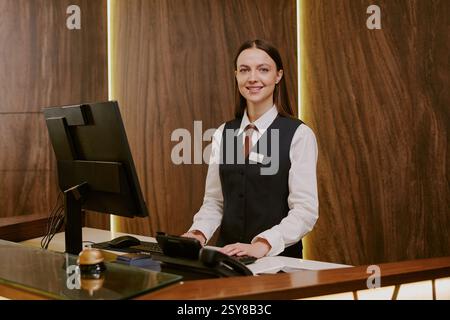 Medium close up of adorable smiling receptionist standing near table with computer while working at hotel Stock Photo