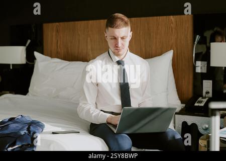 Medium close up of young businessman just dropped his jacket on bed working on laptop after hotel check-in Stock Photo