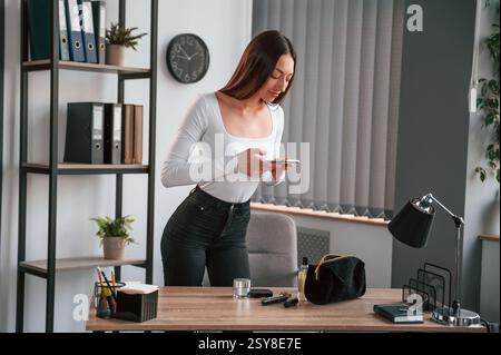 Standing and using laptop. Young beautiful woman in white clothes is in the office. Stock Photo