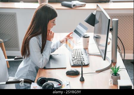 SIde view. Sitting by computer. Young female designer is in the modern office. Stock Photo