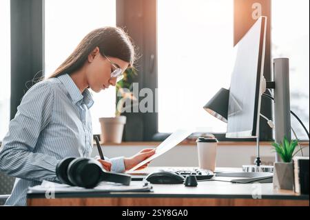 SIde view. Sitting by computer. Young female designer is in the modern office. Stock Photo