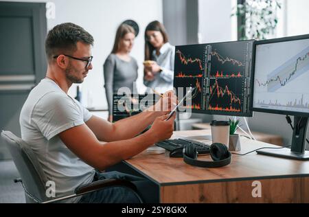 Stock broker is sitting by computer and working with exchange technology. Women is standing and talking behind. Stock Photo