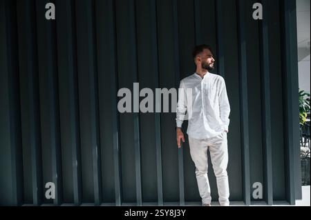 Young handsome man with beard and in white shirt is standing against dark green background. Stock Photo