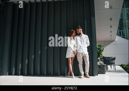 Near the modern building. Beautiful young couple is standing together against dark green background. Stock Photo