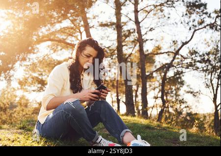 Using smartphone. Beautiful woman is sitting on the grass in the forest at daytime. Stock Photo