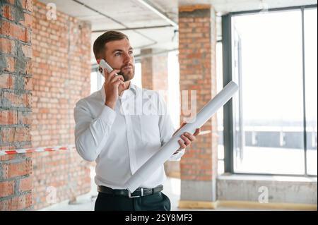 Talking by phone. Man in white shirt is working indoors at construction site. Managing the project. Stock Photo