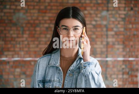 Talking by phone. Young woman is standing in the unfinished building on construction site and working on a project. Stock Photo