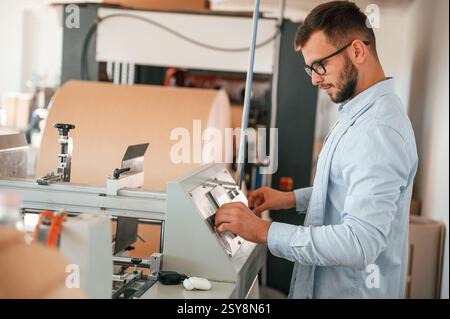 Operating printing cutting machine. Print house worker in white clothes is indoors. Stock Photo