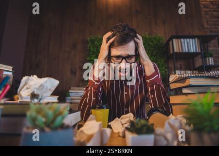 Stressed young man overwhelmed with work and studies in a cozy home setting surrounded by books and crumpled papers Stock Photo