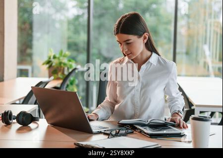 With laptop. Beautiful young woman in white clothes is working in office. Stock Photo