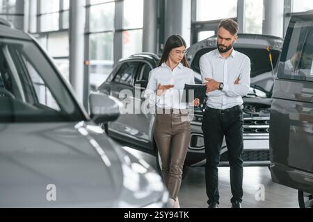 Reading information by using tablet. Man with woman in white clothes are in the car dealership together. Stock Photo