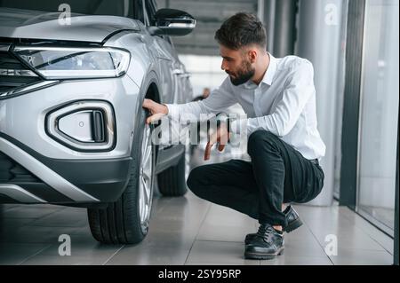 Checking the tires. Young man in white clothes is in the car dealership. Stock Photo