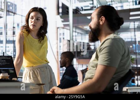 Happy coworkers discussing holiday plans during break in office. Employee enjoying downtime at work, chatting with friendly colleague, idling in workspace waiting for computer update Stock Photo