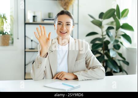 Positive charming business woman waving her hand while working in an office setting, greeting someone over video call, enjoying her virtual meeting in a friendly atmosphere, conducting brainstorm Stock Photo