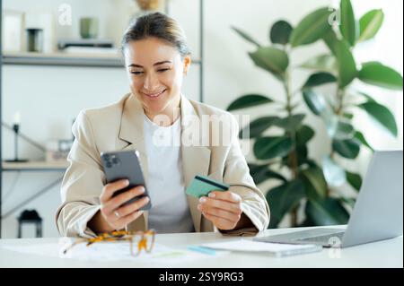 Confident successful business woman, smiling and holding a credit card and smartphone in hands while sitting at a desk in her modern office, completing online tasks, doing online payments Stock Photo
