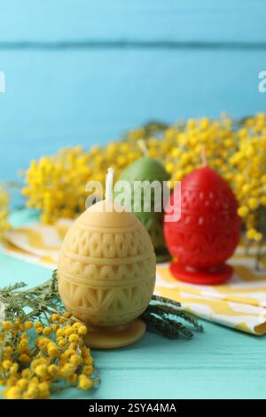 Colorful egg-shaped candles and mimosa flowers on turquoise wooden table against light blue background, closeup. Easter decor Stock Photo