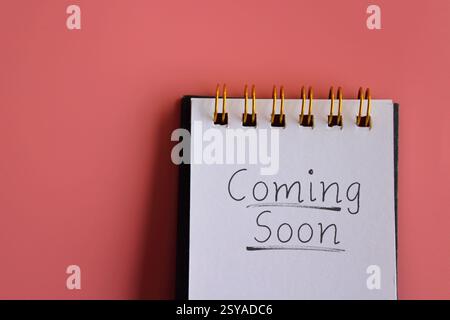 A notepad with the handwritten phrase "Coming Soon" on a bright pink background. Copy space for text. Announcement, reveal, teaser concept. Stock Photo