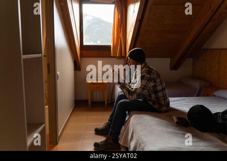 A man sits on a bed in a cozy cabin, engrossed in his smartphone. The room has a rustic wooden interior, enhanced by natural light from a window showi Stock Photo