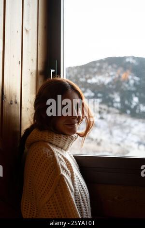 A young woman with red hair wearing a cozy, white knitted sweater stands by a wooden window, gazing at a snowy mountain landscape, embodying a peacefu Stock Photo