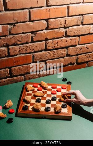 A creative take on checkers using chicken nuggets as game pieces on a board, set against a rustic brick wall. A hand engaged in play, bringing a fun t Stock Photo