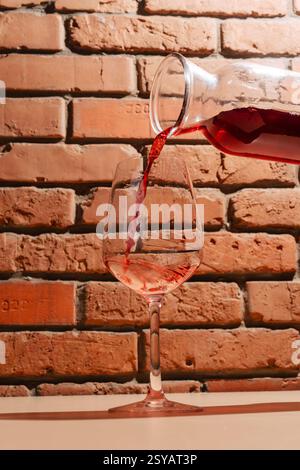 A stream of red wine flows from a glass carafe into a wine glass, set against a rustic brick wall. The warm tones highlight the rich color and texture Stock Photo