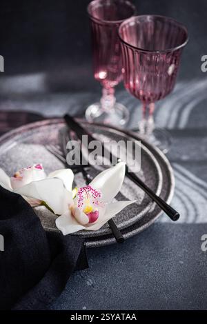 A sophisticated table setting featuring textured plates, dark cutlery, and delicate white orchids. Two pink glasses enhance the romantic ambiance agai Stock Photo