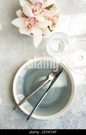 Stylish table setting features a textured ceramic plate, shiny cutlery, and delicate orchid flowers on a soft gray surface, creating a serene and eleg Stock Photo
