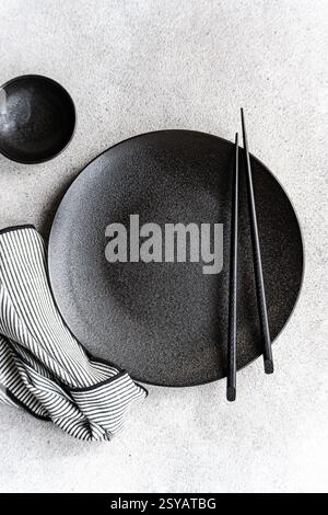 A minimalist black and white table setting featuring a textured dark plate, matching bowl, pair of chopsticks, and a striped cloth on a light backgrou Stock Photo