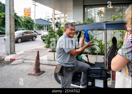 A cheerful Thai tuk tuk driver in a gray shirt is seen interacting with unrecognizable tourists, engaging in conversation, and maintaining his vehicle Stock Photo