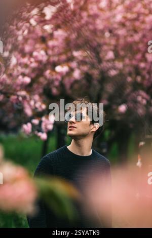A young man wearing round sunglasses enjoys the vibrant scenery of lush cherry blossoms and colorful tulips in bloom. The soft focus on the blooming t Stock Photo