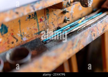 A set of well-used paintbrushes rests on a vintage wooden easel covered in colorful paint splatters. The close-up shot captures the textures and artis Stock Photo