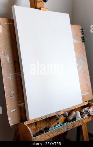 A clean, blank canvas rests on a well-used wooden easel covered in paint splatters. Art supplies sit nearby, waiting for the next creative masterpiece Stock Photo