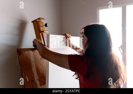 A young woman paints a canvas in her home studio, capturing a moment of artistic creativity. Sunlight filters through the window, highlighting her foc Stock Photo