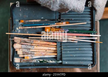 A set of well-worn paintbrushes, covered in dried paint, rests in a fabric case on an artist’s table. The assortment of brushes reflects the creativit Stock Photo