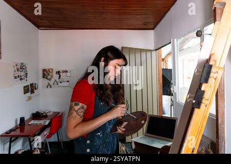 A Latin young woman is focused on painting a canvas in her cozy home art studio. She holds a palette and brush, surrounded by artistic tools and inspi Stock Photo