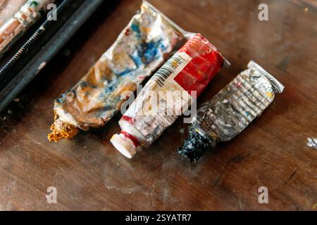 A close-up of worn and crumpled paint tubes covered in dried paint, resting on a wooden table. The messy textures and vibrant colors highlight the cre Stock Photo