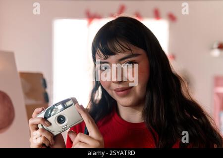 A young woman with long dark hair and a warm smile holds a vintage film camera. She wears a red shirt in a creative studio setting, exuding confidence Stock Photo