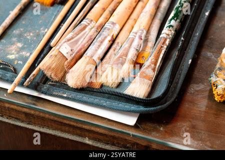 A close-up of well-used paintbrushes with dried paint resting in a fabric case on a wooden table. The worn textures and colorful bristles reflect the Stock Photo
