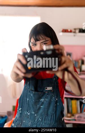 A young woman in a red shirt and paint-splattered overalls playfully holds a vintage camera, aiming it at the viewer. The creative setting and nostalg Stock Photo