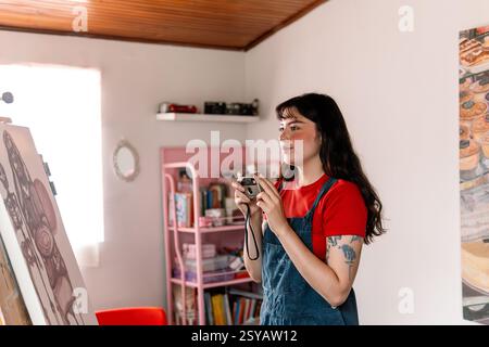 A young woman in a red shirt and paint-splattered overalls holds a vintage camera while standing in her creative studio. Shelves with books, cameras, Stock Photo