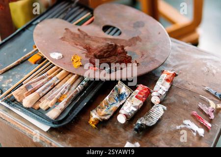 A wooden palette with mixed colors rests on a case of paintbrushes, with used paint tubes nearby. The scene highlights the artistic process and the to Stock Photo