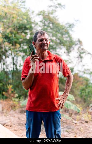 Elderly Colombian man in a red shirt using a smartphone outdoors, standing amidst a rural setting with trees in the background. His expression shows e Stock Photo