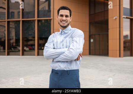 A young professional in business casual attire stands confidently with arms crossed, smiling in front of a modern office building. His positive expres Stock Photo
