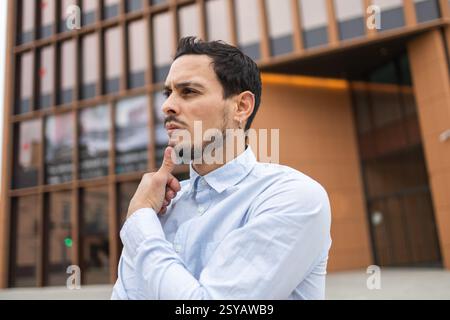 A young man in a light blue shirt stands in front of a modern glass building, touching his chin in a thoughtful pose. His serious expression conveys d Stock Photo