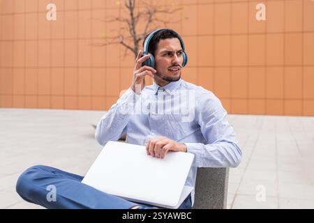 Man in business casual attire sits outside with a laptop and headphones, appearing focused while listening. The modern urban setting emphasizes produc Stock Photo