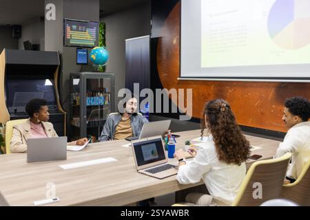 A diverse team collaborates in a modern office setting. The group is engaged in a presentation with laptops, notes, and a projector screen. Stock Photo