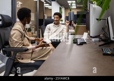 Two colleagues converse in a modern office setting, sitting across from each other. The environment is professional yet relaxed, with plants adding a Stock Photo
