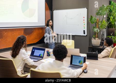 A team engaged in a dynamic office meeting. A presenter explains data on a whiteboard, with attentive colleagues taking notes on laptops, fostering co Stock Photo